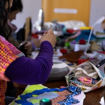 Manos trabajando sobre una arpillera de colores, con figuras azules y rosadas.