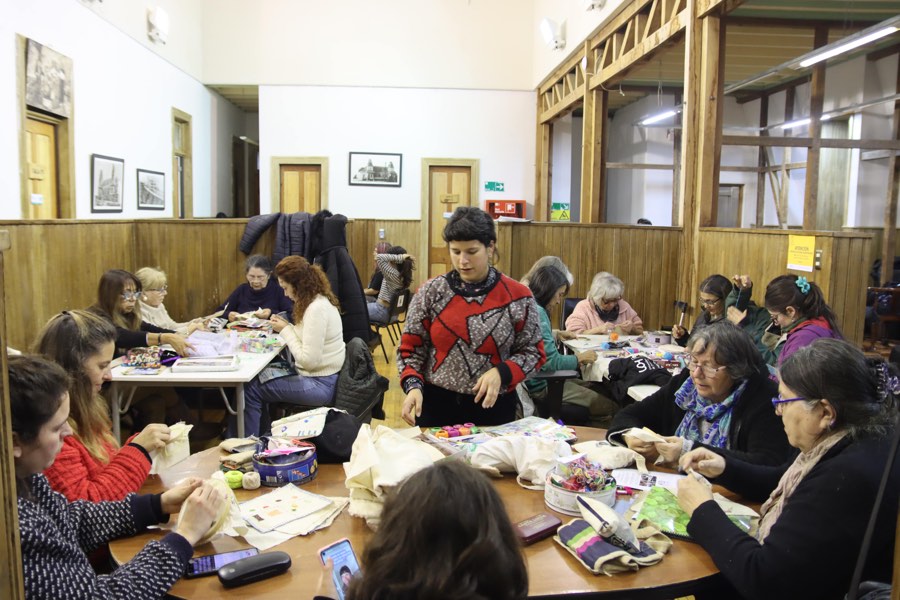 Una mesa larga rodeada de mujeres bordando sobre retazos de tela, en un salón de piso y muros de madera.