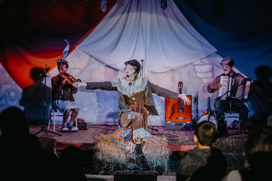 Los tres payasos de Circoliflor en escena: uno al centro con los brazos abiertos, otro toca el violín y el tercero el acordeón, ante un telón de tela y con público sentado delante.