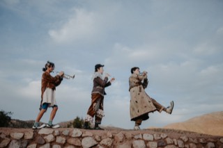 Los tres caminan en fila sobre un muro de piedra tocando trompeta, flauta y otro instrumento, con el cielo y los cerros detrás.