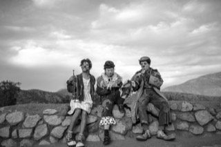 Los tres sentados en un muro de piedra al atardecer, con sus instrumentos, en una fotografía en blanco y negro.