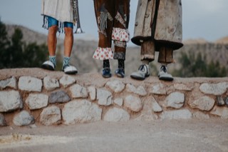 Detalle de los seis zapatos de los tres payasos, de pie sobre un muro de piedra, con calcetines de colores y pantalones remendados.