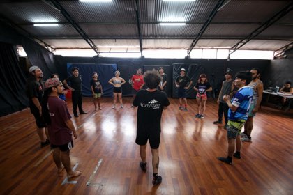 Una rueda de participantes de pie en una sala de ensayo, escuchando al profesor que está en el centro.