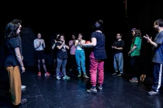 Una rueda de participantes aplaudiendo en un escenario oscuro mientras la profesora, en el centro, sostiene una pelota roja.